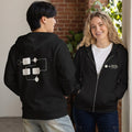 Two people wearing black hoodies with logos, standing indoors near a brick wall.